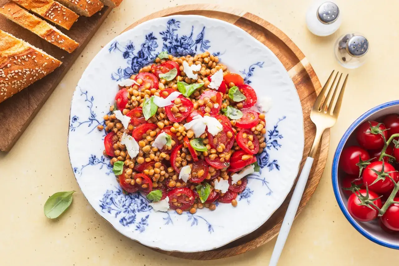 Un'insalata di lenticchie colorata con pomodorini, basilico e scaglie di formaggio, servita su un piatto decorato. Accanto, pane a fette e una ciotola di pomodorini freschi.