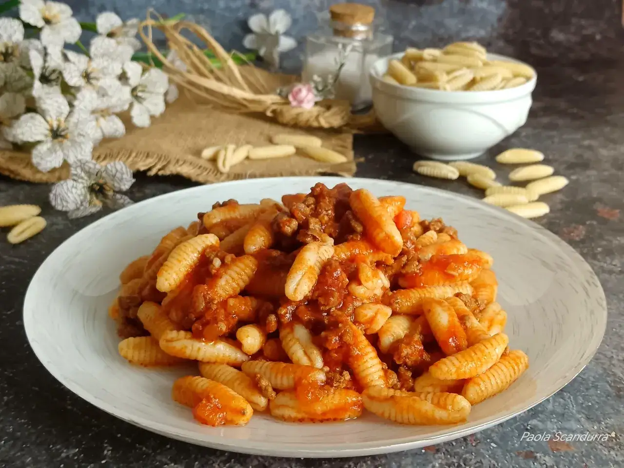 Piatto di malloreddus alla campidanese, pasta sarda con rag&ugrave; di salsiccia e pomodoro, decorato con fiori bianchi e spighe.
