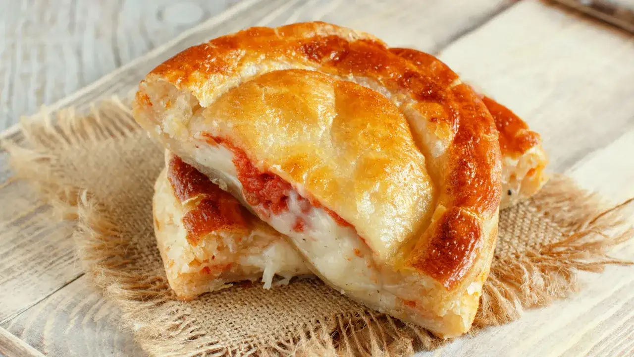 Rustici con pasta sfoglia ripieni di formaggio filante e pomodoro, dorati e invitanti su un tagliere di legno.