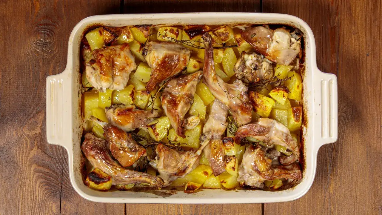 Delizioso coniglio al forno con patate dorate e rosmarino, servito in una teglia bianca su un tavolo di legno.