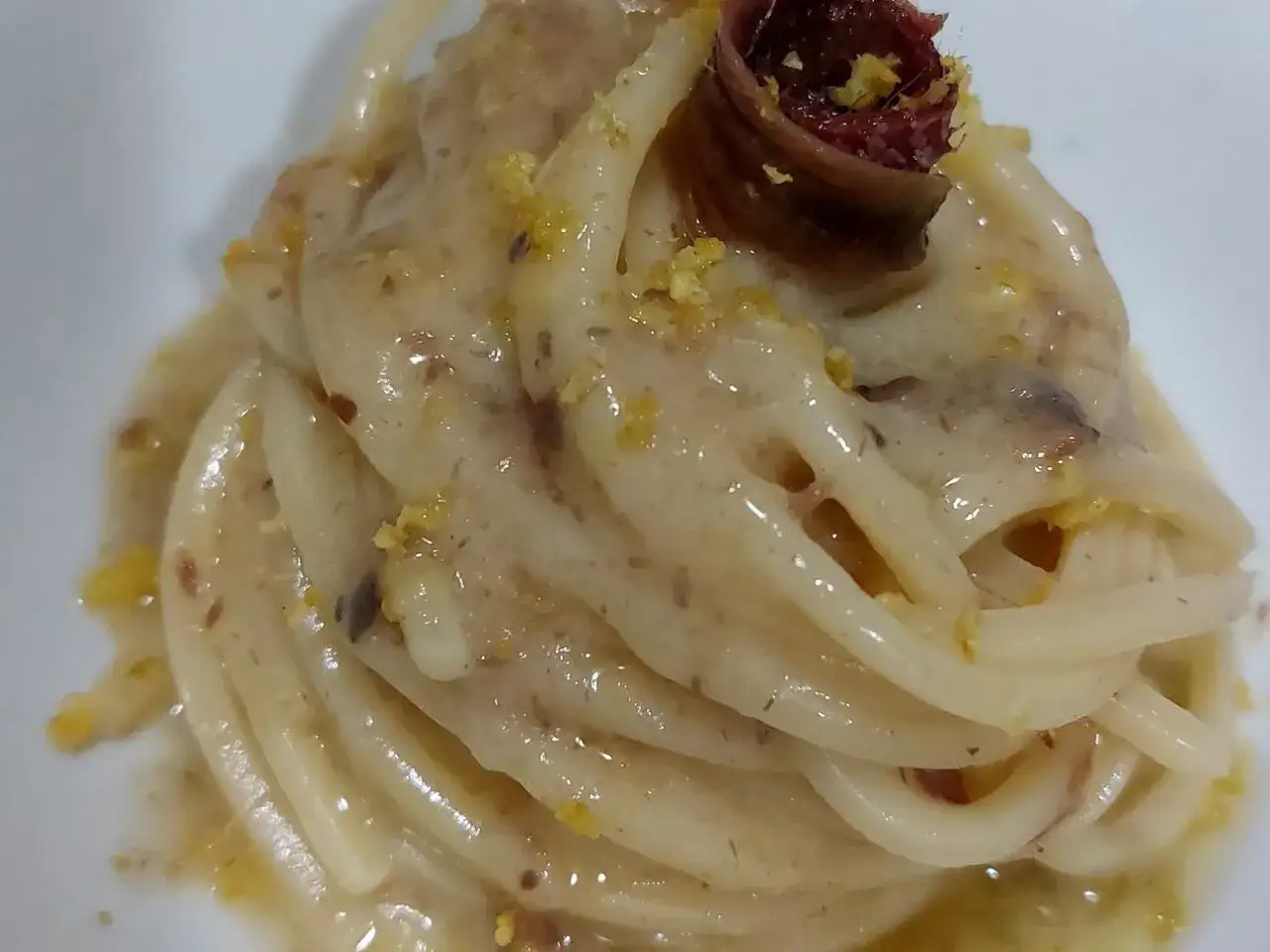Un piatto di pasta burro e alici, condita con una salsa cremosa e guarnita con scorza di limone e un'alice arrotolata.
