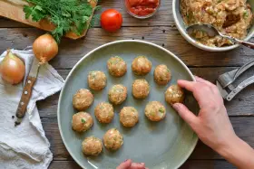 Polpette di pollo pronte per essere cucinate, disposte su un piatto grigio, con ingredienti freschi come cipolle e pomodoro sullo sfondo.