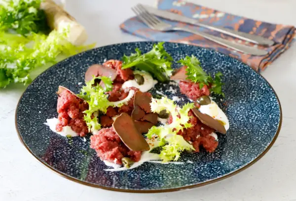 Deliziosa tartare di manzo con lattuga croccante, capperi e una crema bianca su un piatto blu decorato.