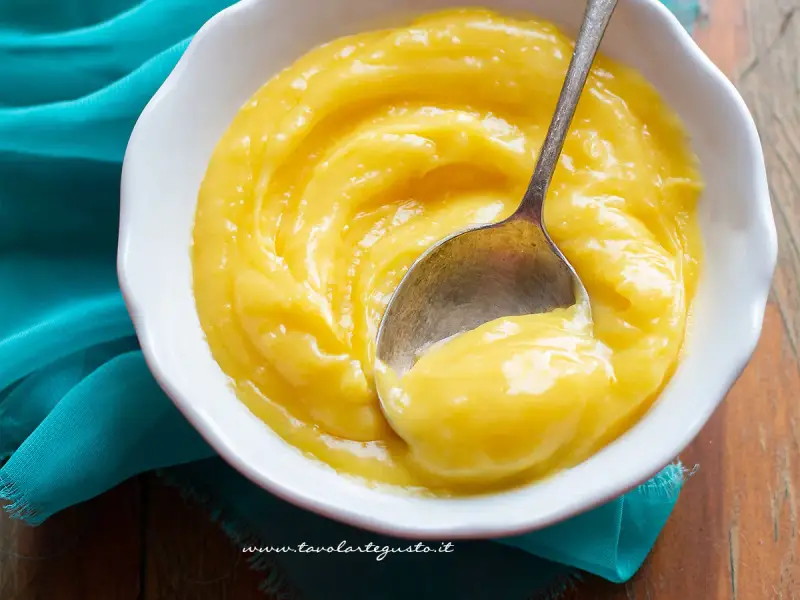 Una ciotola bianca piena di vellutata crema al limone, con un cucchiaino che ne raccoglie una porzione.